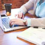 A person sitting in front of a laptop at a desk with a textbook to their left and a smartphone and cup of tea to their right.