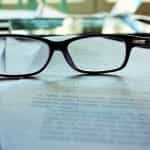 Papers on a desk with black-framed eyeglasses on top