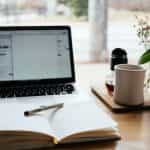 An open notebook lying flat on a table in front of a laptop next to a coffee mug and flower vase.