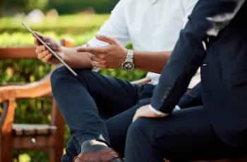 Two people in business wear sitting on a bench outside and discussing matters together, with one person showing information to the other on a tablet device.